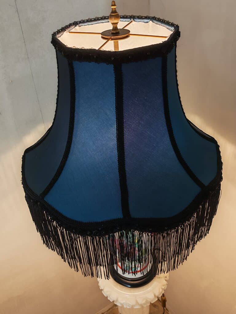 Abat-jour Bleu Vintage à Franges – Éclairage Décoratif Chic Abat-jour bleu évasé avec franges noires décoratives et tresses, sur une lampe vintage allumée. Éclairage chaleureux.