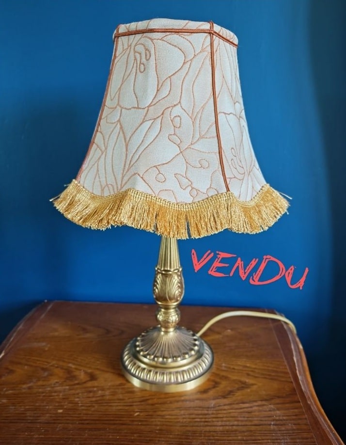 Lampe de table vintage en laiton ouvragé avec abat-jour floral beige et orange, franges dorées, sur table en bois. Le texte VENDU est visible.