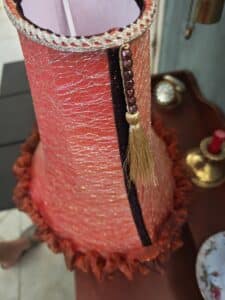 Abat-jour rouge orangé style corset, filet scintillant doré, bordure tressée argentée, perles, pompon doré. Sur une table avec objets.