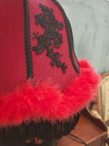 Abat-jour rouge et noir avec maille résille, dentelle florale noire, bordure en plumes rouges vives et franges noires. Ambiance boudoir élégante.