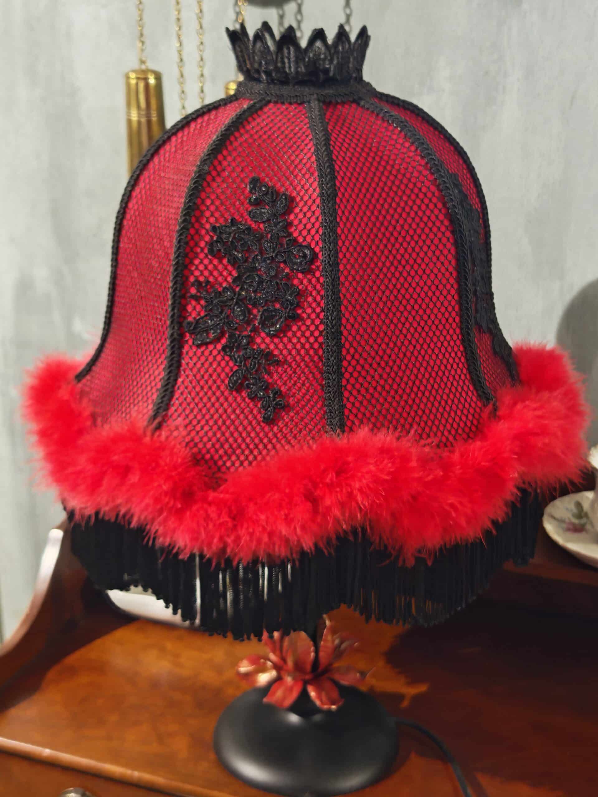 Abat-jour décoratif rouge et noir, style boudoir, avec motif dentelle, plumes et franges. Couronne noire, sur table en bois.