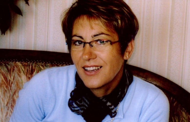 Portrait souriant : femme aux lunettes et pull bleu Femme souriante aux cheveux courts bruns, portant des lunettes, un pull bleu clair et une écharpe foncée. Assise sur un canapé.