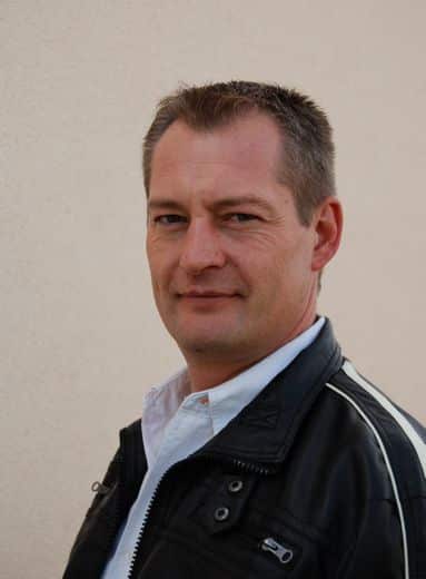 Homme en blouson cuir : style décontracté et confiant Homme d'âge moyen souriant, blouson cuir noir aux rayures blanches sur les bras, chemise blanche, devant un mur clair.