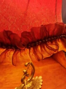 Lampe décorative rouge orangé avec tissu froissé doré, bordure plissée de perles foncées et ornement doré.