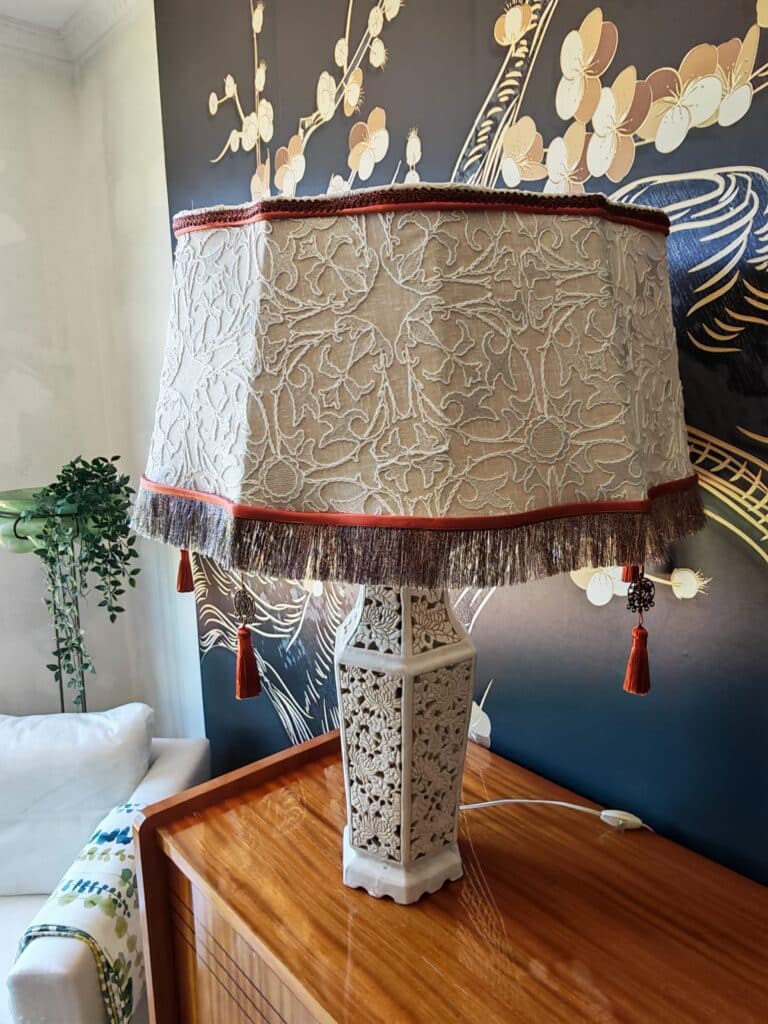 Lampe de table beige, abat-jour brodé à franges et pompons rouges, base sculptée florale sur bois. Fond mural sombre motifs orientaux.