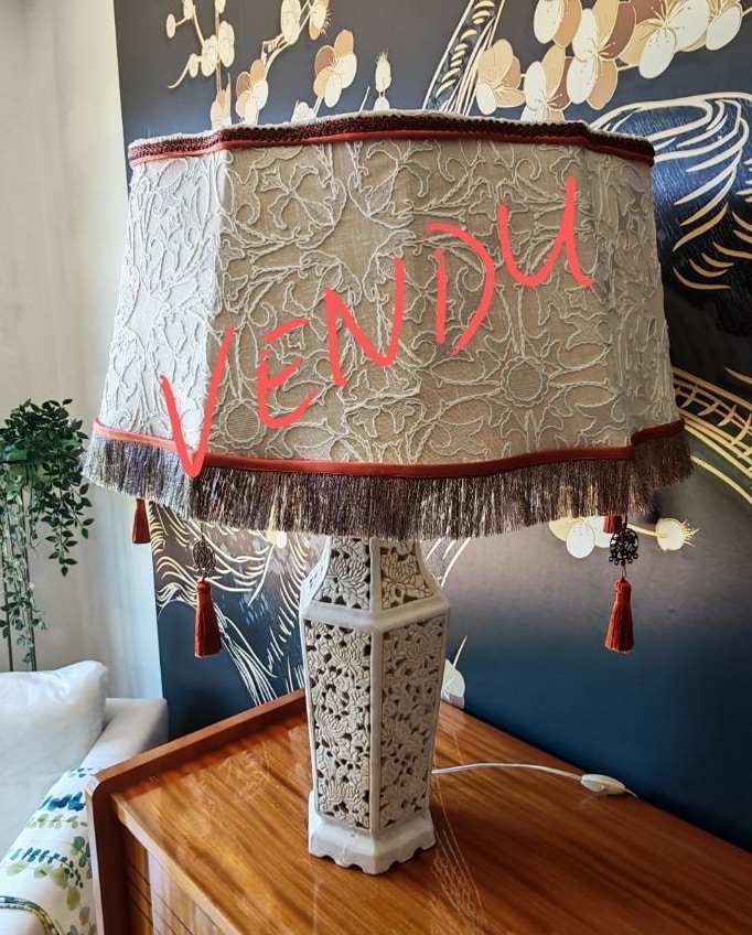 Grande lampe de table VENDUE avec abat-jour octogonal brodé de motifs floraux, franges et pompons rouges. Base sculptée crème, sur meuble en bois foncé, devant un mur décoré de motifs asiatiques.