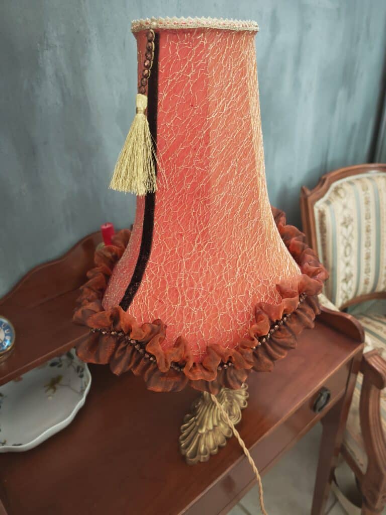 Lampe de table vintage avec abat-jour corail à motifs dorés, gland, perles et volants, posée sur une table en bois foncé. Mur bleu-gris en fond.