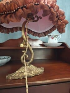Lampe de table vintage en laiton avec abat-jour marron à volants et perles, posée sur un meuble en bois foncé près d'une tasse de thé fleurie.