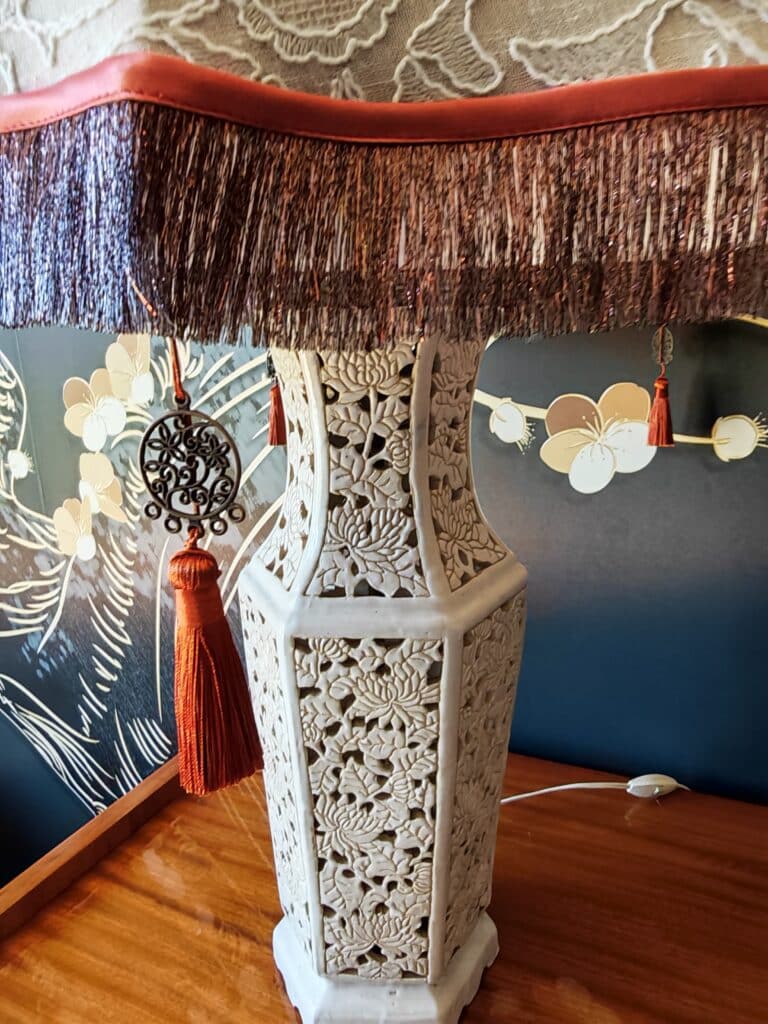 Élégante lampe de table orientale blanche en forme de vase sculpté de motifs floraux, abat-jour frangé rouge-brun, sur une table en bois poli.