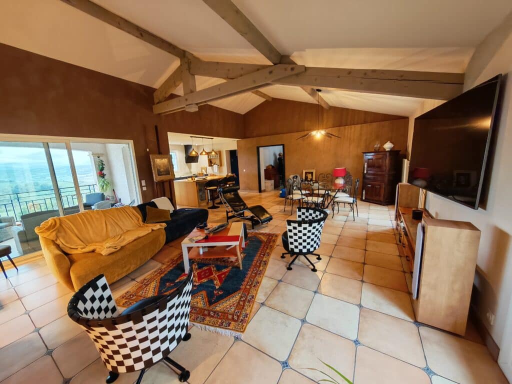 Salon rustique-moderne avec poutres apparentes, sol carrelé, canapé jaune, et vue sur une vallée verdoyante.