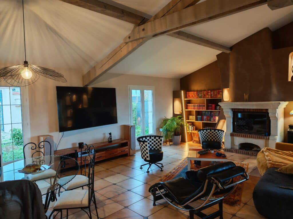 Salon chaleureux avec poutres apparentes, télévision grand écran, fauteuils design (dont un LC4 noir), cheminée en pierre et étagères remplies de livres.