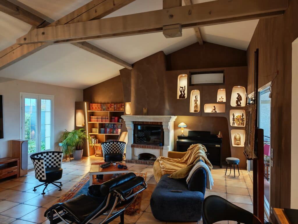 Salon chaleureux avec poutres apparentes, cheminée rustique, étagères remplies de livres, sculptures africaines rétroéclairées et fauteuils design.