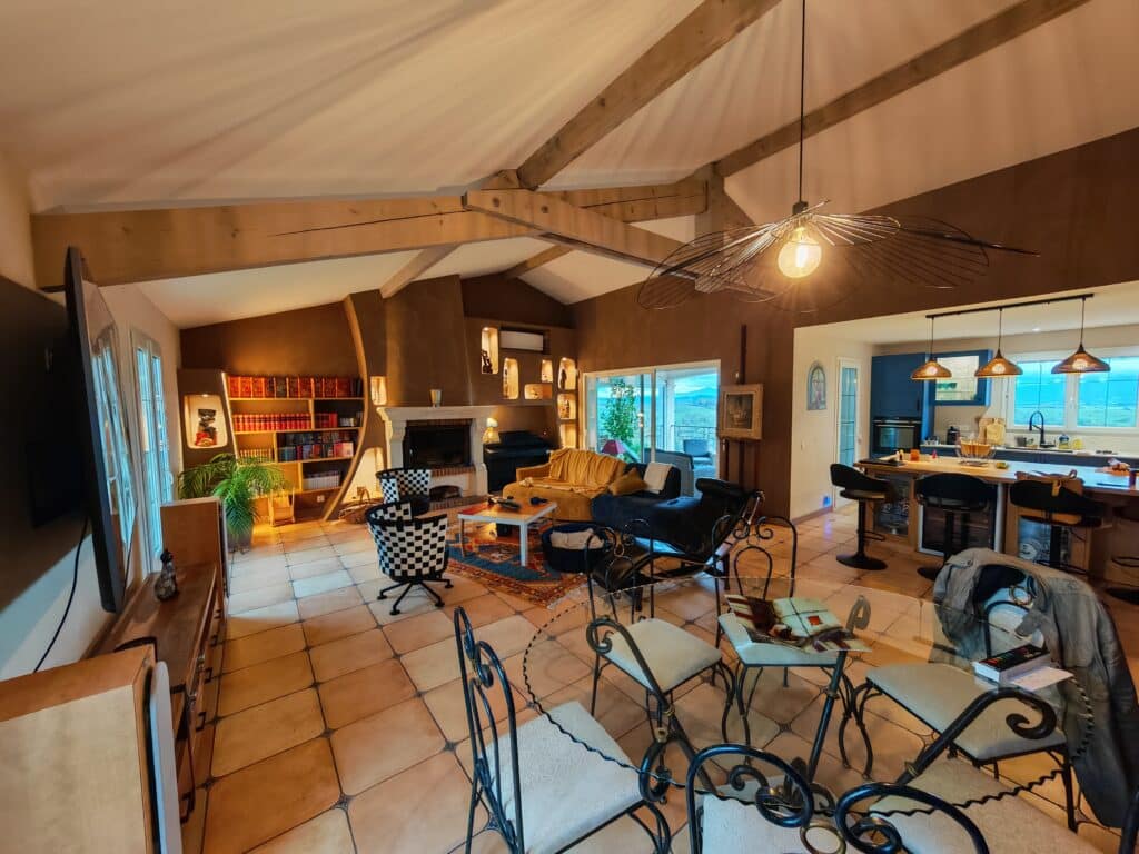 Vaste salon-séjour rustique-chic avec poutres apparentes, cheminée, bibliothèque intégrée et cuisine ouverte avec îlot. Vue sur la nature.