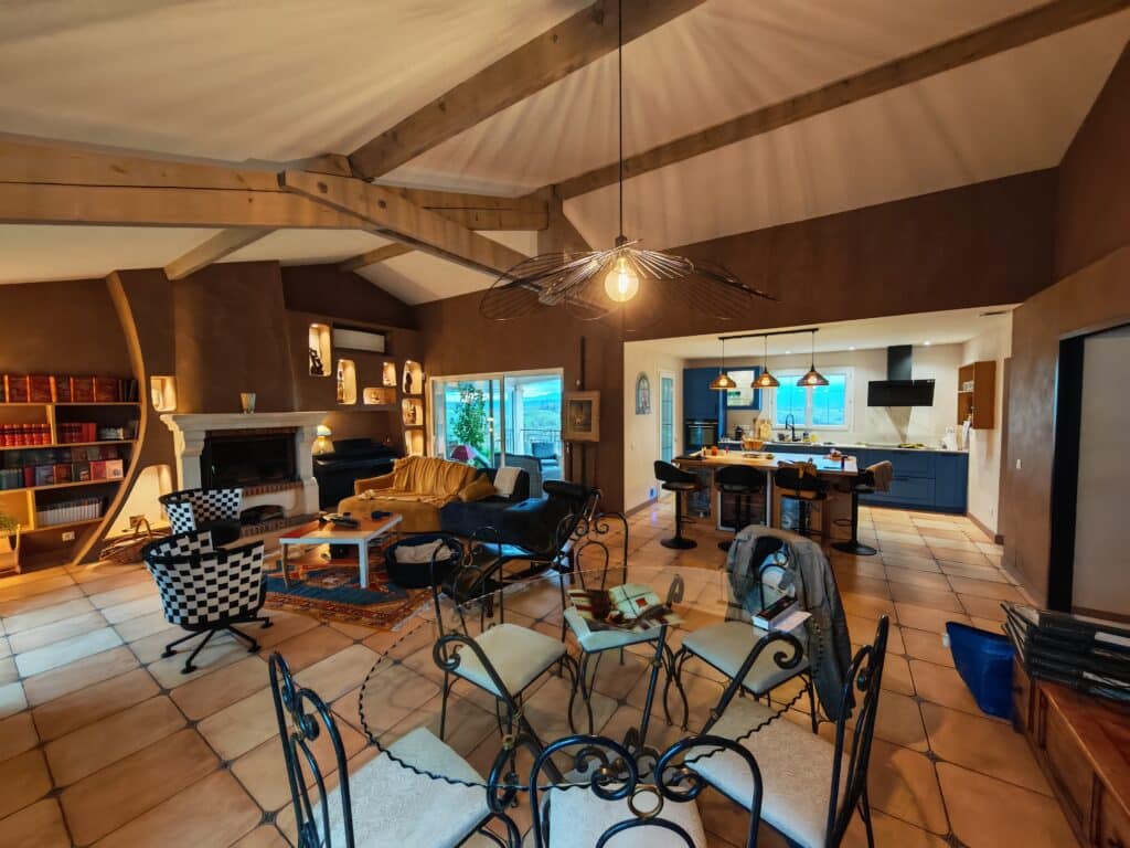 Grand salon rustique-chic avec poutres apparentes, mobilier confortable, cheminée, étagères encastrées, et cuisine ouverte avec îlot central.