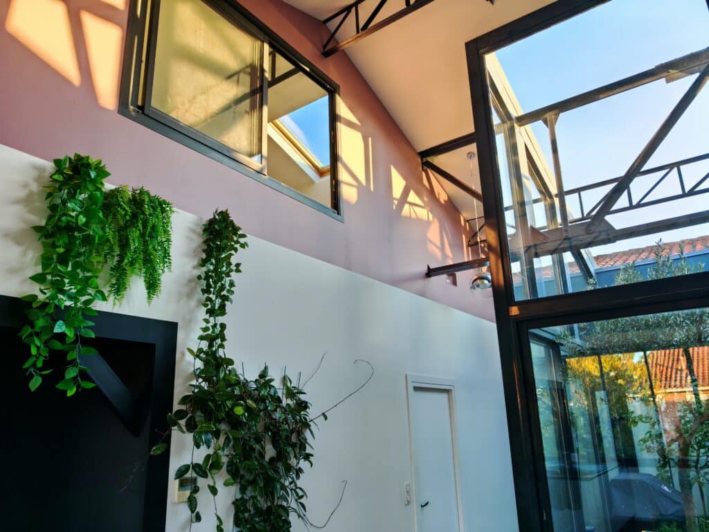 Design intérieur moderne: mur bicolore rose/blanc, verrière atelier noire et poutres métalliques. Lumière naturelle et plantes vertes suspendues.