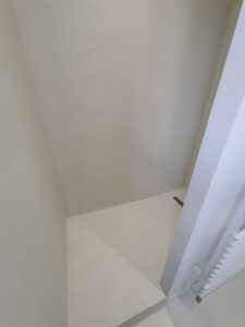 Coin de pièce aux murs grège neutre. Un radiateur vertical blanc borde l'espace étroit. Un petit drain sombre est visible au sol.