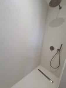 Douche italienne minimaliste avec murs en béton ciré clair. Robinetterie et douchette gris mat moderne. Drain linéaire.