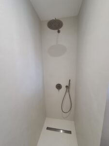 Douche moderne minimaliste en microciment blanc. Pommeau de pluie, mitigeur encastré gris mat et caniveau linéaire discret.