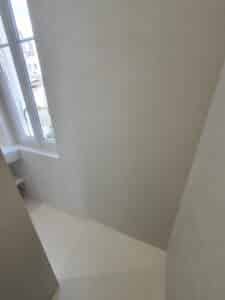 Intérieur moderne aux murs et sol en micro-ciment gris-beige uniforme. Lumière naturelle intense par la fenêtre de toit blanche.