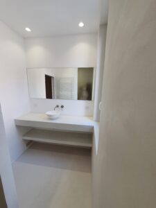 Salle d'eau minimaliste: vasque sur plan flottant et étagère en microciment clair. Grand miroir mural, éclairage par spots.