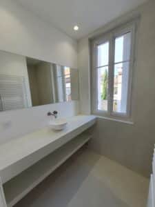 Salle de bain moderne monochrome. Plan vasque flottant en microciment clair, miroir mural, robinetterie inox et fenêtre lumineuse.